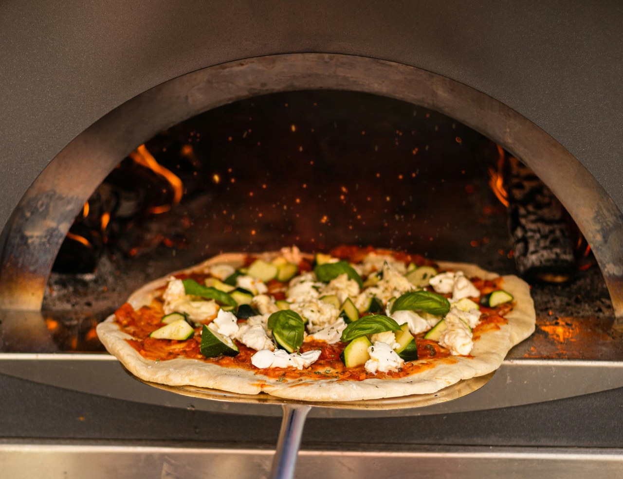 A pizza topped with mozzarella, courgette and basil being slid into a wood-fired oven